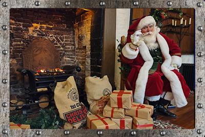 Santa sitting on a chair next to a fireplace and lots of presents wrapped in brown paper and tied up with red ribbon on the floor